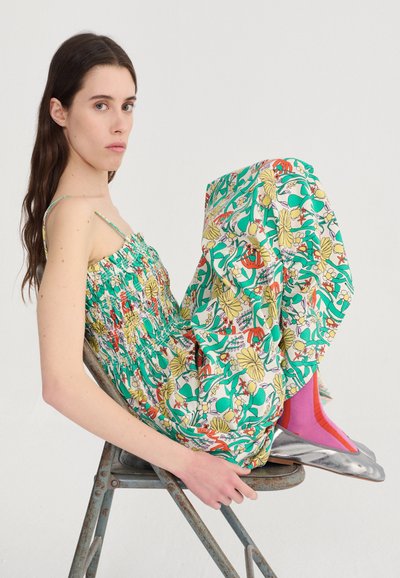 Jonge vrouw met lang donker haar zit zijwaarts op een metalen stoel, gekleed in een kleurrijke bloemenjurk, roze sokken en zilveren platte schoenen tegen een effen witte achtergrond.