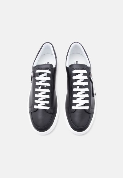 Paire de baskets basses noires avec des lacets blancs et des semelles blanches, vues de dessus sur un fond gris clair.