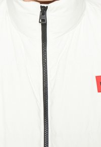 Weiße gepolsterte Jacke mit hohem Kragen, schwarzem Reißverschluss und einem kleinen roten Logopatch auf der linken Brust. Glatte Textur und minimalistisches Design.