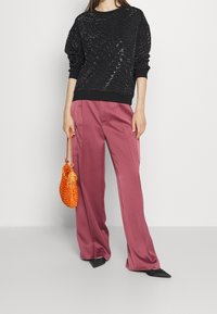 Suéter negro texturizado con cuello redondo, combinado con pantalones sueltos rosas. La modelo sostiene un bolso de crochet naranja y lleva unos zapatos de punta negros.