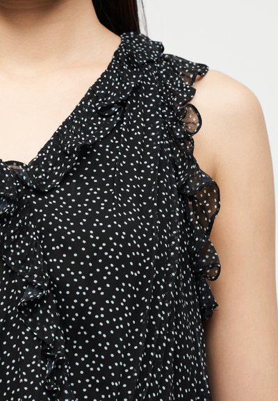 Κοντινό πλάνο του ώμου μιας γυναίκας που φοράει μαύρο αμάνικο φόρεμα με λευκές πουά και βολάν στα άκρα.