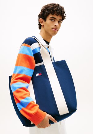 Joven con un suéter de rayas naranja y azul que lleva un bolso tipo tote grande azul marino con asas blancas y un pequeño parche con logo.