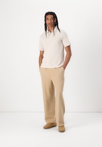 Polo beige a maniche corte abbinato a pantaloni larghi color sabbia chiara. Il modello indossa sneakers color sabbia; trame di tessuto morbido e casual.