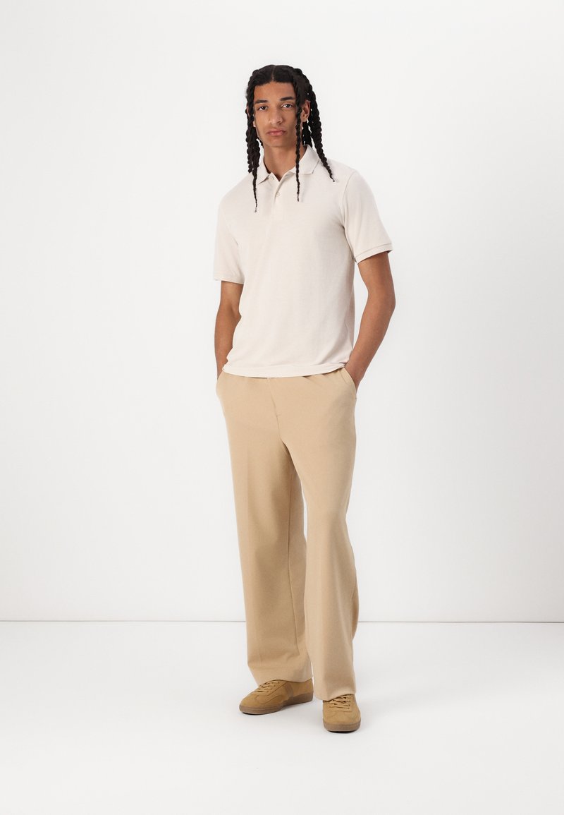 Polo beige a maniche corte abbinato a pantaloni larghi color sabbia chiara. Il modello indossa sneakers color sabbia; trame di tessuto morbido e casual.