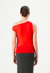Mujer con un moño trenzado que lleva una blusa roja de un solo hombro y una falda texturizada de color gris oscuro, de pie mirando hacia el lado opuesto de la cámara.