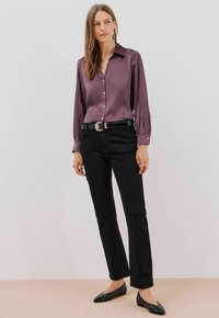 Blusa in satin viola con maniche lunghe e chiusura a bottoni, abbinata a jeans neri a gamba dritta e scarpe nere con borchie in argento. Cintura con fibbia metallica.