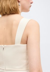 Νεαρό άτομο με κοντά ξανθά μαλλιά, που φοράει ένα αμάνικο κρεμ φόρεμα με φερμουάρ στην πλάτη και ασημένιο σκουλαρίκι σε σχήμα σταγόνας, σε λευκό φόντο.