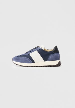 Tummansininen mokkanahkainen sneakerit, joissa on valkoiset raidat, pyöreä kärki, nauhalla suljettava muotoilu ja ruskea kumiulkopohja. Varressa on kontrastina musta kantapää.