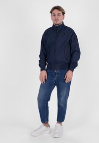 Giubbotto bomber blu navy con chiusura a zip, polsini e orlo a coste, due tasche frontali. Paio di jeans azzurri e sneakers bianche.
