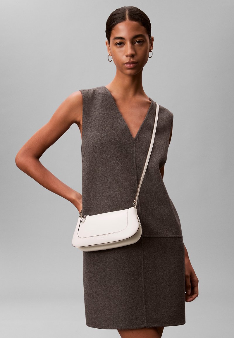 Femme aux cheveux lisses portant une robe marron sans manches et un sac bandoulière blanc, sur fond gris uni.