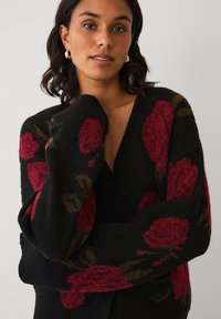 Zwarte cardigan met groot rood rozenpatroon, voorzien van een V-hals en knoopsluiting. Zachte textuur en een losse pasvorm met lange mouwen.