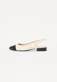 Chaussure slingback avec un dessus crème et un embout noir brillant. Elle présente un petit talon carré et des découpes sur les côtés.