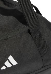 Bolsa de deporte negra hecha de tela duradera, con una superficie texturizada, una cremallera sobresaliente y un destacado diseño del logo blanco de Adidas.