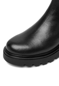 Čierna kožená topánka s hrubou, robustnou gumovou podrážkou a textúrovaným povrchom, umiestnená na bielom pozadí.