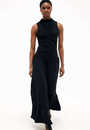 Vrouw met kort haar, gekleed in een mouwloze zwart gerimpelde top en wijde zwarte broek, loopt naar voren op een witte achtergrond.