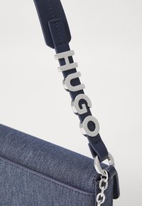 Mørkeblå denimveske med en glatt lærreim. Sølvfargede "HUGO"-bokstaver som fremhever reimen, med metallbeslag og kjededetaljer.