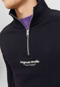 Černá mikina na zip s vysokým límcem, se stříbrným zipem a bílým textem "Originals Studio" na přední straně. Materiál z bavlněné směsi.
