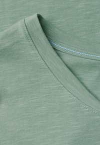 T-shirt in cotone verde con scollo a V, tessuto texturizzato e dettaglio di cucitura blu lungo il colletto.