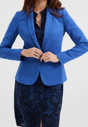 Blue tailored blazer with a notch lapel, solid color, and long sleeves, worn over a dark floral patterned dress. Smooth texture, fitted design.