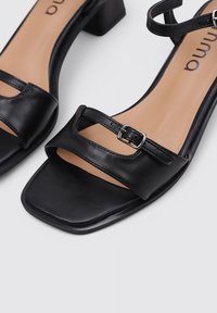 Sandalias de tacón de cuero negro con puntas cuadradas, que presentan dos correas y una hebilla para ajuste. Plantilla suave en tono marrón claro y tacón bloque resistente.