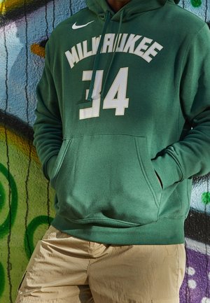 Person iført en grøn Milwaukee hættetrøje med nummer 34 og beige bukser, der står op ad en graffiti-væg med hænderne i hættetrøjer.