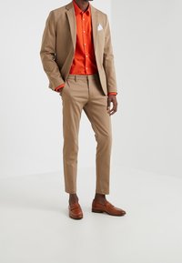 Man in een beige pak en bruine loafers, draagt een feloranje overhemd met een witte zakdoek in de zak van zijn jas, staat met zijn hand in zijn zak.