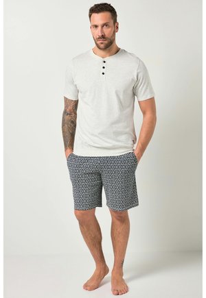 Mand med tatoveret arm iført en lysegrå kortærmet henley-shirt og sort-hvide shorts med geometrisk mønster, stående barfodet.