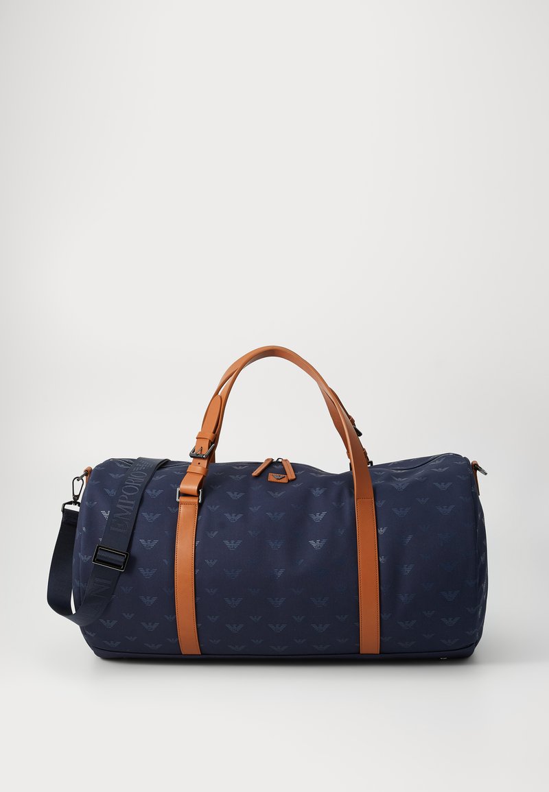 Saco de duffle azul-marinho com padrão de logotipo em relevo, detalhes em pele castanha, alças duplas e uma correia de ombro ajustável. Textura suave.