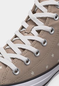 Κοντινό πλάνο μπεζ αθλητικού παπουτσιού με λευκά αστέρια και χοντρές λευκές κορδόνια που περνούν μέσα από ασημένιες τρύπες.