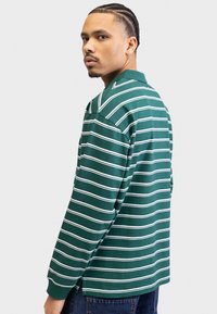Green long-sleeve polo shirt featuring horizontal white stripes, a collared neckline, and side slits at the hem. Worn with dark denim jeans.