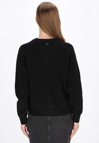 Schwarzer Strickpullover mit gerippter Textur, Raglanärmeln und rundem Halsausschnitt. Besitzt ein kleines Logo-Tag auf der Rückseite.