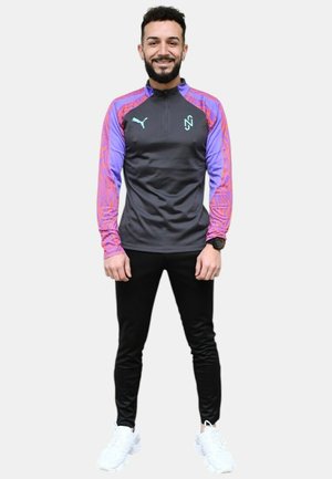 Chaqueta deportiva Puma con cuerpo negro, mangas moradas con un patrón de olas, cuello con cremallera, y pantalones deportivos negros a juego con zapatillas blancas.