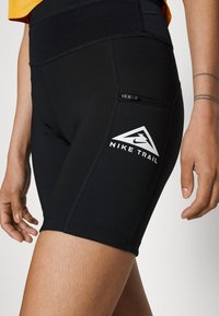 Gros plan sur un short de sport noir Nike Trail avec une petite poche zippée sur la cuisse, porté par une personne avec des tatouages visibles sur le bras.