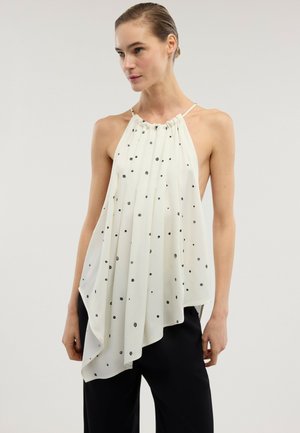 Femme portant un haut dos nu blanc ample à pois noirs avec un ourlet asymétrique, associé à un pantalon noir, sur un fond gris uni.