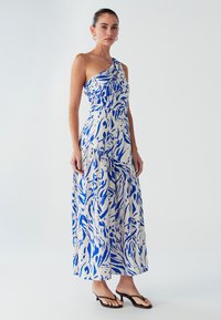 Robe maxi asymétrique crème avec motifs abstraits bleus, corsage froncé et jupe fluide. Associée à des tongs noires.