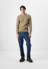 Pull en maille beige à côtes avec des poignets et un ourlet côtelés, associé à un jean en denim bleu et des baskets noires, se tenant contre un fond clair.