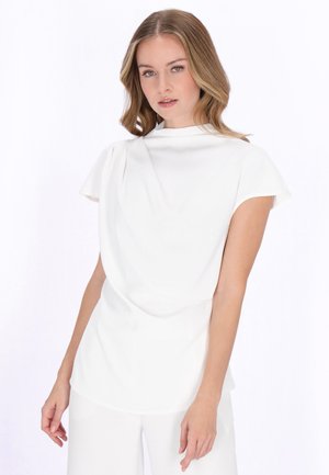 Młoda kobieta z blond włosami do ramion, ubrana w biały krótki rękaw i pasujące białe spodnie na jednolitym tle.