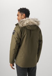Maslinasto zelena parka jakna s umjetnim krznom na kapuljači, crni logotip na rukavu i glatka, bezteksturna površina.
