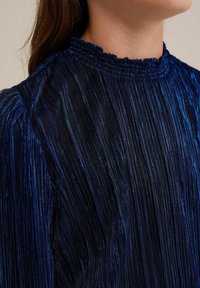 Blå plisserad blus med hög, smockad krage. Har vertikala glittrande ränder och mjukt, texturerat tyg med en elegant fall.