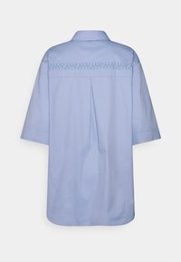 Camisa de algodão azul claro com mangas curtas, colarinho e pregas nas costas. Apresenta bordados decorativos ao longo da parte superior das costas, acrescentando detalhe.