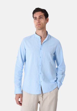 Uomo con capelli scuri che indossa una camicia azzurro chiaro a maniche lunghe con bottoni e pantaloni beige, in piedi con una mano in tasca, guardando leggermente a destra.