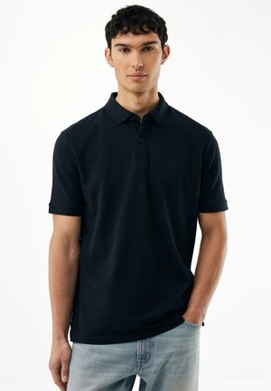 Giovane uomo con capelli corti e ricci, indossa una polo nera e jeans azzurri, in piedi con una mano in tasca.