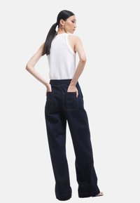 Tummansiniset korkeavyötäröiset farkut, joissa on kaksi takataskua ja näkyvät saumat, yhdistettynä valkoiseen hihattomaan toppiin, jossa on neuleyksityiskohta.