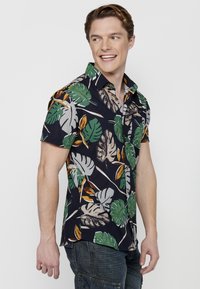 Camisa de manga corta con fondo azul marino y grandes hojas de monstera verdes, acentos en naranja y marrón, y un diseño de cuello clásico.