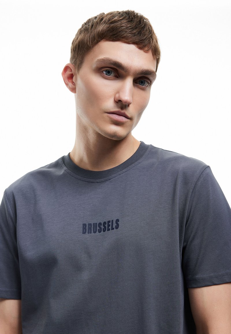 Graues Rundhals-T-Shirt aus weichem Stoff mit strukturierter Oberfläche. Mit "BRUSSELS" in Dunkelblau quer über die Brust bedruckt.