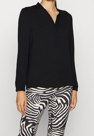 Vrouw draagt een zwarte blouse met lange mouwen en een V-hals en een zwart-witte zebra-print broek, staat met handen ontspannen langs haar lichaam.