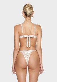 Lingerieset van satijnen stof in lichtbeige. De top heeft verstelbare bandjes met een strikdetail op de rug, en de onderkant is in de vorm van een string.