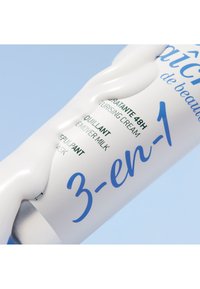 Vit tub av 3-i-1 återfuktande kräm med blå text; slät textur med en böjd, konturerad form. Bakgrunden är ljusblå.