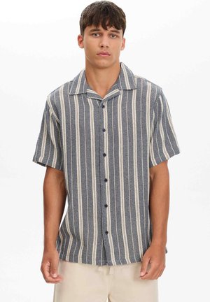 Joven con una camisa de manga corta abotonada con rayas verticales azul marino y blancas y pantalones de color claro, de pie contra un fondo blanco.