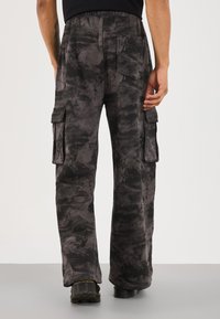 Rear view of person wearing black and gray camouflage cargo pants with six pockets and black shoes, standing on a gray floor.